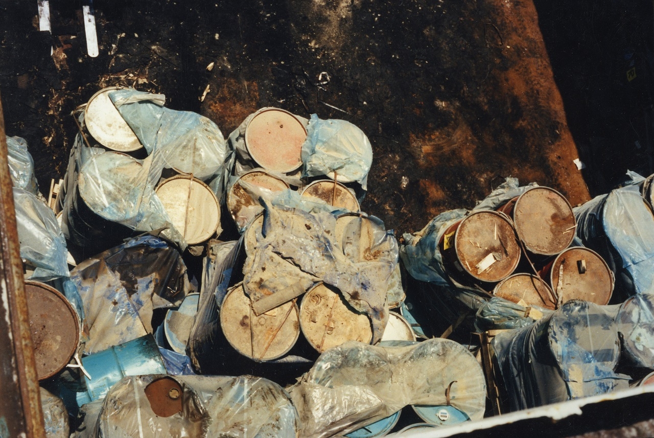 Rusty-looking barrels are grouped in fours, partially-covered in plastic tarp, and tied together.