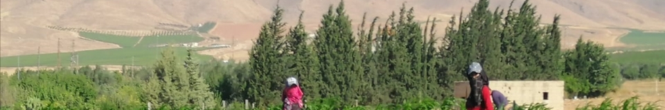 Two women in colorful long dresses, gloves, face-covering scarves, and hats look towards the camera as they stand in a lush field against a mountainous backdrop.